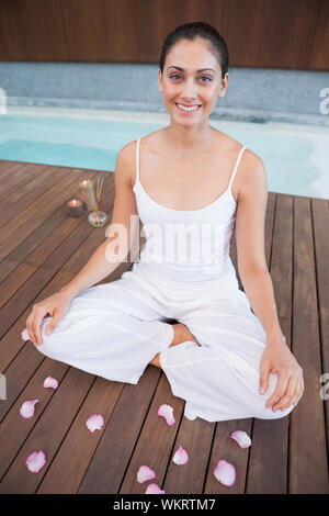 Friedliche Brünette in weißen Kleidern sitzen im Lotus Pose von Blütenblättern in Health Spa Umgeben Stockfoto