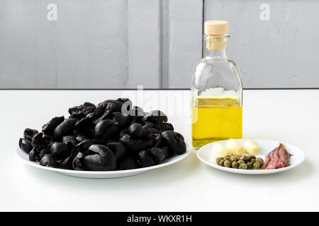 Apenade Zutaten: Entkernten schwarzen Oliven auf weiße Platte, Olivenöl in Glas Flasche, drei Knoblauchzehen, einige Anchovisfilets und eingelegte Kapern. Stockfoto