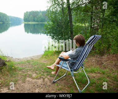 Junge sitzt im Liegestuhl ein Buch lesen auf einem See Stockfoto