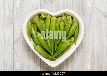 Frische organische Grün okra in Herzform Schüssel auf ein weißes Holzbrett isoliert. Stockfoto