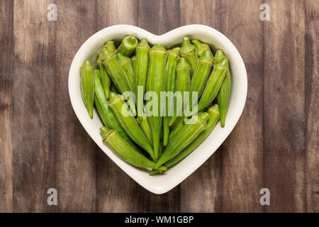 Frische organische Grün okra in Herzform Schüssel auf eine rote Holzbrett isoliert. Stockfoto