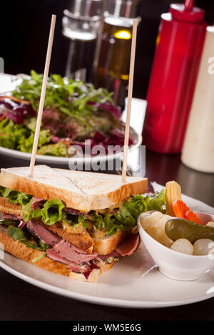 Köstliche dünn geschnittenes Fleisch pastrami Club Sandwich mit frischen curly Kopfsalat mit einer kleinen Schale von eingelegtem Gemüse auf einem weißen Teller serviert, isoliert Stockfoto