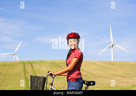 Nachhaltige Zukunft Konzept. Mädchen auf dem Fahrrad mit Feld- und Windkraftanlagen. Blauer Himmel mit kopieren. Schöne Gemischte asiatische/kaukasisch-Modell. Stockfoto