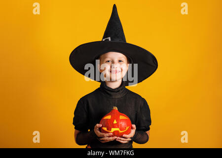 Süße kleine Mädchen in eine Hexe Kostüm hält einen Kürbis jack Lantern auf gelbem Hintergrund. Halloween Feier. Stockfoto