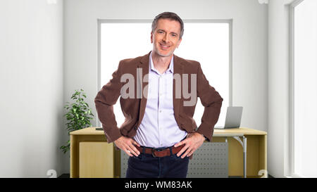 Middle-Aged Geschäftsmann mit grauem Haar und trug eine Braune Jacke stehend in einem Büro. Zeigt beim Systemstart und Corporate Business. Er lächelt und h Stockfoto