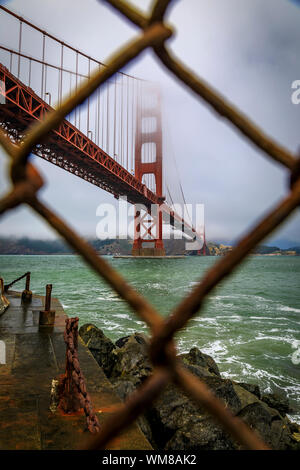 Die berühmte Golden Gate Bridge gesehen durch ein rostiges Maschendrahtzaun an einem bewölkten Sommertag mit niedrig hängenden Nebel rollen in San Francisco, Kalifornien Stockfoto