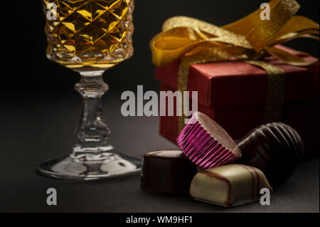 Nahaufnahme eines Clusters von köstlichen handgemachten edle Pralinen mit einem Becher Weißwein und ein Geschenk mit goldenen Bogen in ein Konzept von einem romantischen annive Stockfoto