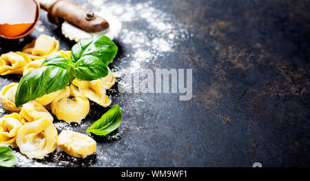 Draufsicht auf hausgemachte Tortellini mit Mehl, Tomate und Basilikum auf dunklen vintage Hintergrund Stockfoto