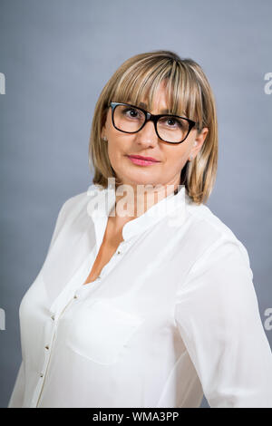 Wissenschaftliche attraktiven Frau mittleren Alters blond in Gläsern Stand mit verschränkten Armen vor einem grauen Hintergrund in die Kamera schaut Stockfoto