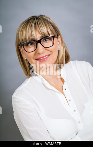 Wissenschaftliche attraktiven Frau mittleren Alters blond in Gläsern Stand mit verschränkten Armen vor einem grauen Hintergrund in die Kamera schaut Stockfoto