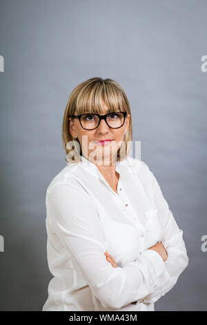 Wissenschaftliche attraktiven Frau mittleren Alters blond in Gläsern Stand mit verschränkten Armen vor einem grauen Hintergrund in die Kamera schaut Stockfoto