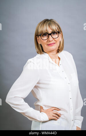 Wissenschaftliche attraktiven Frau mittleren Alters blond in Gläsern Stand mit verschränkten Armen vor einem grauen Hintergrund in die Kamera schaut Stockfoto