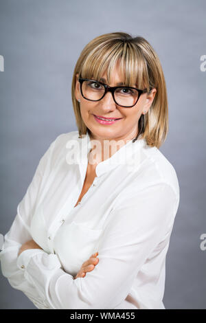 Wissenschaftliche attraktiven Frau mittleren Alters blond in Gläsern Stand mit verschränkten Armen vor einem grauen Hintergrund in die Kamera schaut Stockfoto