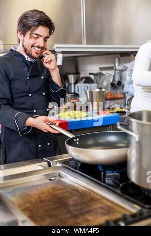 Professionelle Koch oder Küchenchef in Uniform mit einem Edelstahl Schüssel in der Hand, einen Anruf auf seinem Smartphone während das Abendessen vorbereiten Stockfoto