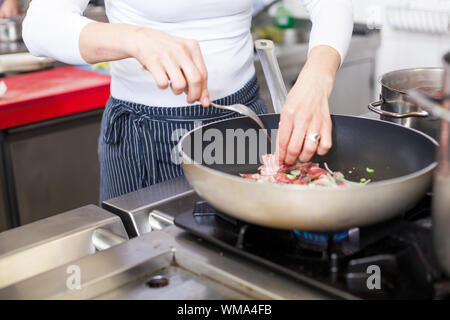 Koch oder Köchin schmoren Fleisch in einer beschichteten Pfanne über eine Gaskochfeld, wie sie eine Mahlzeit in einer gewerblichen Küche bereitet Stockfoto