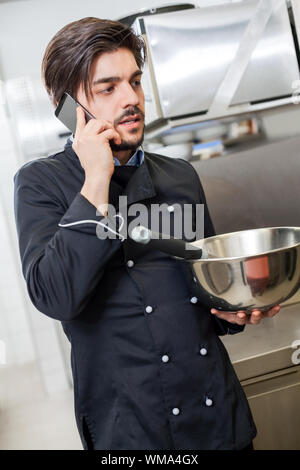 Professionelle Koch oder Küchenchef in Uniform mit einem Edelstahl Schüssel in der Hand, einen Anruf auf seinem Smartphone während das Abendessen vorbereiten Stockfoto