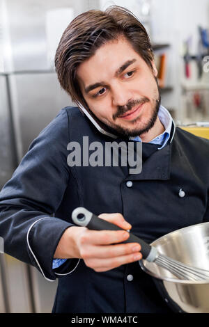 Professionelle Koch oder Küchenchef in Uniform mit einem Edelstahl Schüssel in der Hand, einen Anruf auf seinem Smartphone während das Abendessen vorbereiten Stockfoto