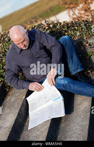 Glatze Mann mittleren Alters in Jeans sitzen im Freien in der Sonne auf einer Treppe eine Zeitung lesen Stockfoto