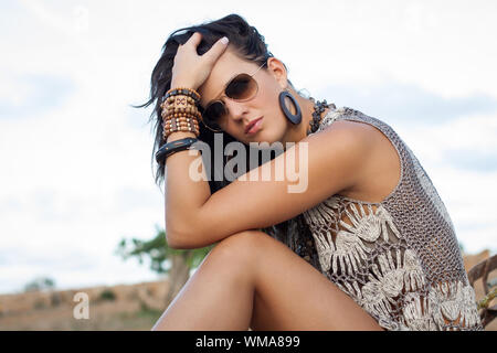 Schöne stilvolle brünette Frau mit Sonnenbrille und mehrere Armreifen auf dem Boden sitzend in der offenen Landschaft seitlich an der Kamera mit der Suche Stockfoto