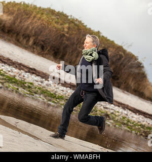 Gerne ältere Frau Herumtollen am Strand schreiten zusammen mit ausgebreiteten Armen und einem Lächeln der Anerkennung, die sie genießt die Natur und die Freiheit ihrer Stockfoto