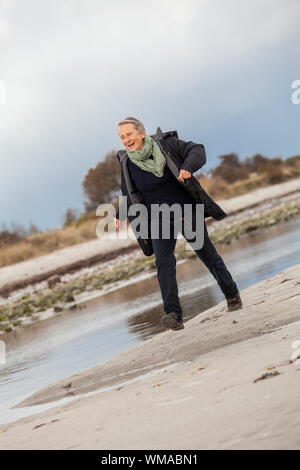 Gerne ältere Frau Herumtollen am Strand schreiten zusammen mit ausgebreiteten Armen und einem Lächeln der Anerkennung, die sie genießt die Natur und die Freiheit ihrer Stockfoto
