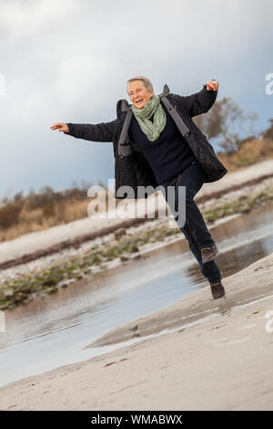 Gerne ältere Frau Herumtollen am Strand schreiten zusammen mit ausgebreiteten Armen und einem Lächeln der Anerkennung, die sie genießt die Natur und die Freiheit ihrer Stockfoto