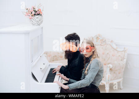 Junges Paar in riesigen Brille sitzt im weißen Raum und spielt Klavier. Stilvolle wulstige Mann und seine blonde Freundin am Flügel sitzen Stockfoto