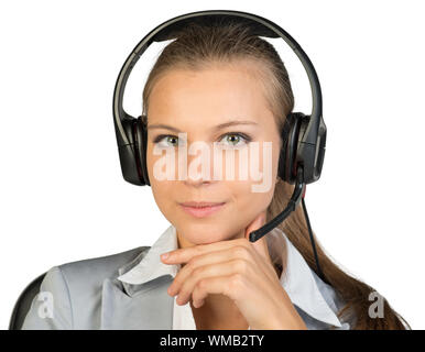 Geschäftsfrau im Headset sitzt auf einem Stuhl, Hand unter das Kinn, an der Kamera schaut. Auf weissem Hintergrund Stockfoto