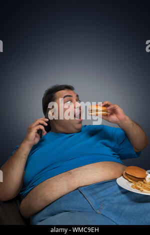 Aufgeregt beleibten Mann mit Bauch sichtbar aus seinem T-Shirt essen Burger beim Gespräch über Mobiltelefon mit einer Platte von Burger und Pommes frites auf seinem Thi Stockfoto