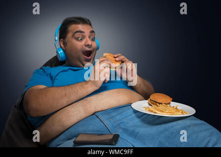 Aufgeregt beleibten Mann mit Bauch sichtbar aus seinem T-Shirt essen Burger, während Sie Musik hören mit einer Platte von Burger und Pommes frites auf seinem Schenkel Stockfoto