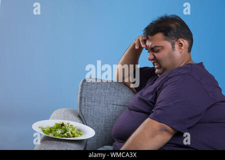 Seitenansicht der beleibten Mann sitzt auf einem Sofa mit der Hand auf die Stirn zeigen Abneigung gegen grünes Gemüse Salat in der Platte Stockfoto