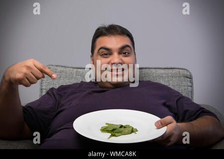 Lächelnd beleibten Mann sitzt auf Stuhl Holding eine Platte mit wenigen Spinatblätter und es gerne zeigen Stockfoto