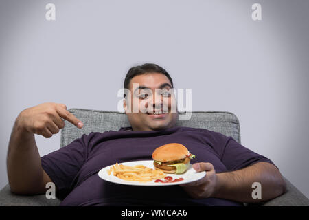 Lächelnd beleibten Mann sitzt auf Stuhl Holding eine Platte mit Burger und Pommes frites an ihm gerne zeigen Stockfoto