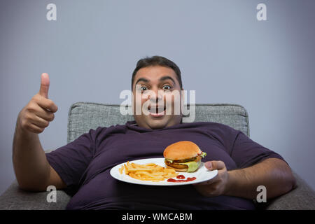 Lächelnd beleibten Mann sitzt auf Stuhl Holding eine Platte mit Burger und Pommes frites, die einen Daumen nach oben anmelden Stockfoto