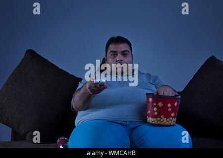 Übergewichtige Menschen zu Hause vor dem Fernseher sitzen auf einem Sofa mit Popcorn und Softdrink Flasche an seiner Seite Stockfoto