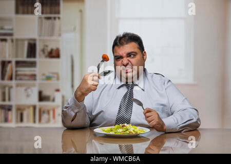 Übergewicht Mann am Esstisch mit Platte voller Gemüse Salat Holding eine Tomate in Scheiben schneiden und mit der Gabel, es mit Abneigung sitzen Stockfoto