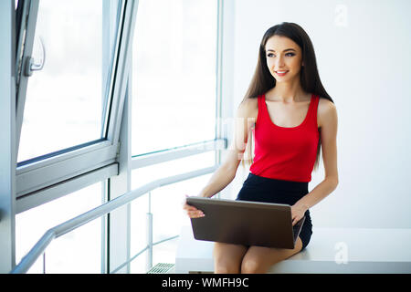 Unternehmen. Schöne junge Frau arbeitet am Computer im Light Modern Office. In Rot T-Shirt und schwarzem Rock gekleidet. Mädchen ist glücklich und lächelnd Stockfoto