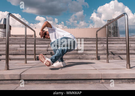 Ein junger Kerl eine Tänzerin, macht eine niedrigere brechen, Tänze in Bewegung im Sommer in der Stadt vor dem Hintergrund der Schritte und ein Gebäude, Jeans Stockfoto