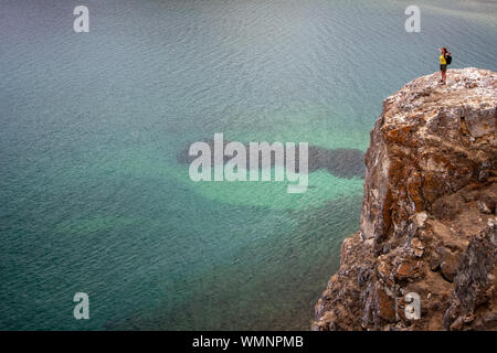 Ein Mädchen mit ausgebreiteten Armen steht auf einem hohen Felsen in der Nähe von Baikal mit grün transparent Wasser. Steile Felsen mit Steinen. Das Wasser ist ruhig. Eine accumulatio Stockfoto