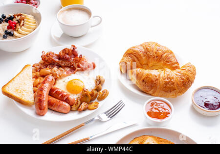 Ansicht von oben flatlay mit Sorten von frischem Frühstück: Spiegeleier mit Speck und Würstchen, Müsli mit Beeren, gebratene Toast mit Marmelade und Butter. Weiß Stockfoto