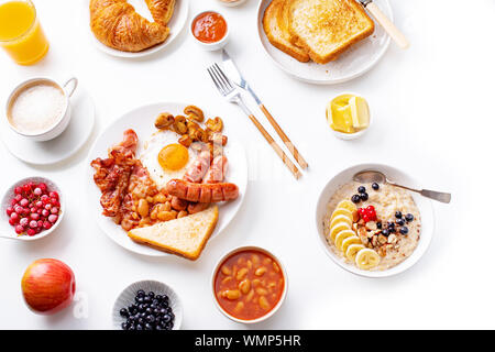 Ansicht von oben flatlay mit Sorten von frischem Frühstück: Spiegeleier mit Speck und Würstchen, Müsli mit Beeren, gebratene Toast mit Marmelade und Butter. Weiß Stockfoto