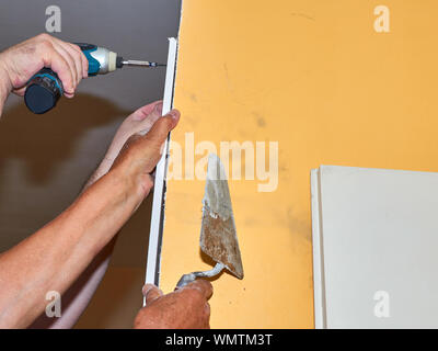 Männliche Hände halten elektrische Schraubendreher und Kelle, während ein Teil der Gipskartonplatten an der Wand während der Renovierungsarbeiten in der Wohnung Stockfoto