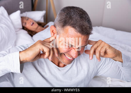 Wütend reifer Mann stecken Ohr mit Finger beim Sitzen in der Nähe von Schnarchen Frau im Bett Stockfoto