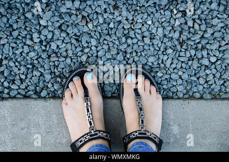 Beine von einem kaukasischen Girl in Sandalen durch einen Kiesweg. Blick von oben. Stockfoto