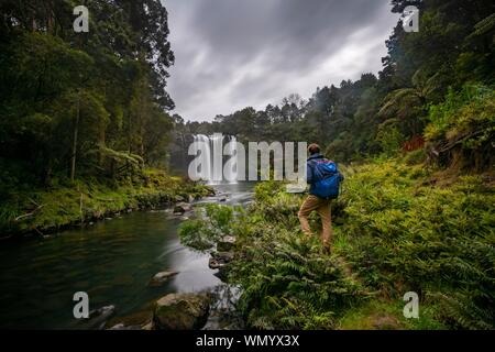 Junger Mann, der vor Wasserfall, Rainbow Falls oder Waianiwaniwa, Kerikeri River, Northland, North Island, Neuseeland Stockfoto