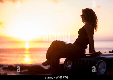 Frau sitzt auf dem Auto mit Strand bei Sonnenuntergang Stockfoto