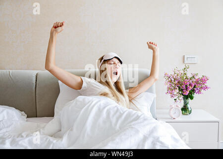 Glückliche junge blonde Frau in Schlafanzug und Augenbinde aufwachen, Gähnen und Strecken im Bett, einen guten Tag. Morgen Licht im Schlafzimmer. Entspannung Stockfoto