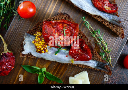 Lecker smorrebrod auf einem Holzbrett. Sandwiches mit schwarzem Roggenbrot, getrocknete Tomaten, gesalzene Sardellen, Senf. Stockfoto