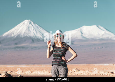 Junge casual blonde Frau allein reisen, kaukasische Mädchen in Hut und Sonnenbrille an Kamera suchen mit schneebedeckten Bergen im Hintergrund. Atemberaubende Stockfoto