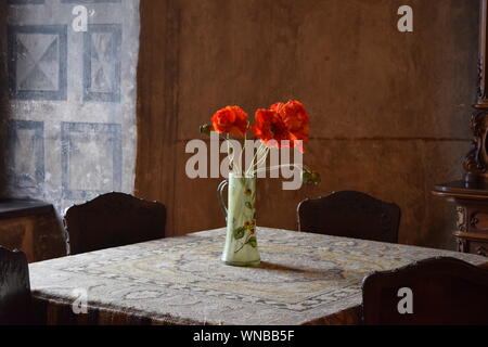 Mohn Blumen in einem Lackiertes Glas Vase, durch helles Sonnenlicht durch ein Fenster beleuchtet, sitzen auf einem Tisch in einem alten Slowakische Burg. Stockfoto